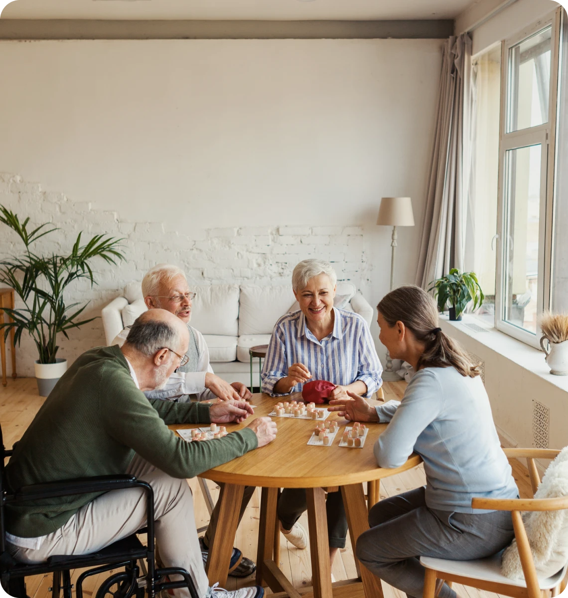 Four elderly people playing cards around a wooden table in a bright room.
